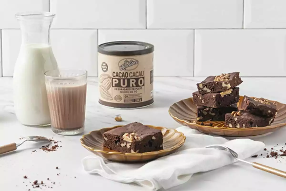 Una gerra de llet al costat d'un got de llet amb cacau en pols i un plat amb brownies de xocolata sobre una taula blanca. Una gerra de llet al costat d'un got de llet amb cacau en pols i un plat amb brownies de xocolata sobre una taula blanca.