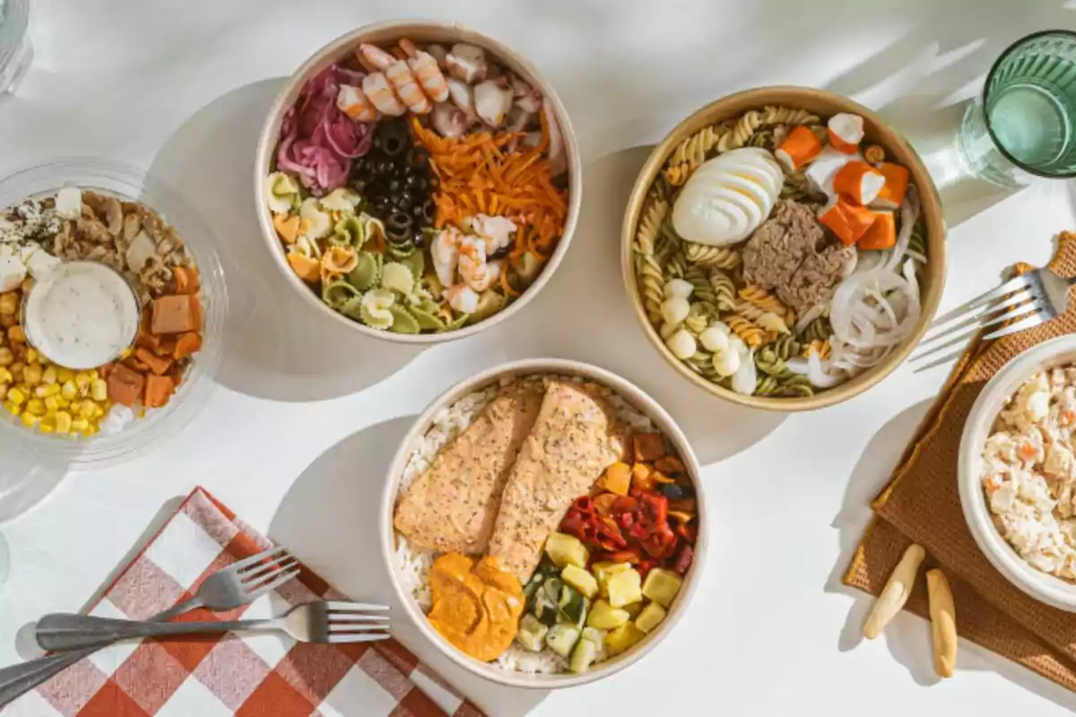 Diverses amanides i bols amb ingredients frescos com pasta, gambes, ou, salmó, verdures i amaniments servits en una taula amb coberts i gots d’aigua Diverses amanides i bols amb ingredients frescos com pasta, gambes, ou, salmó, verdures i amaniments servits en una taula amb coberts i gots d’aigua