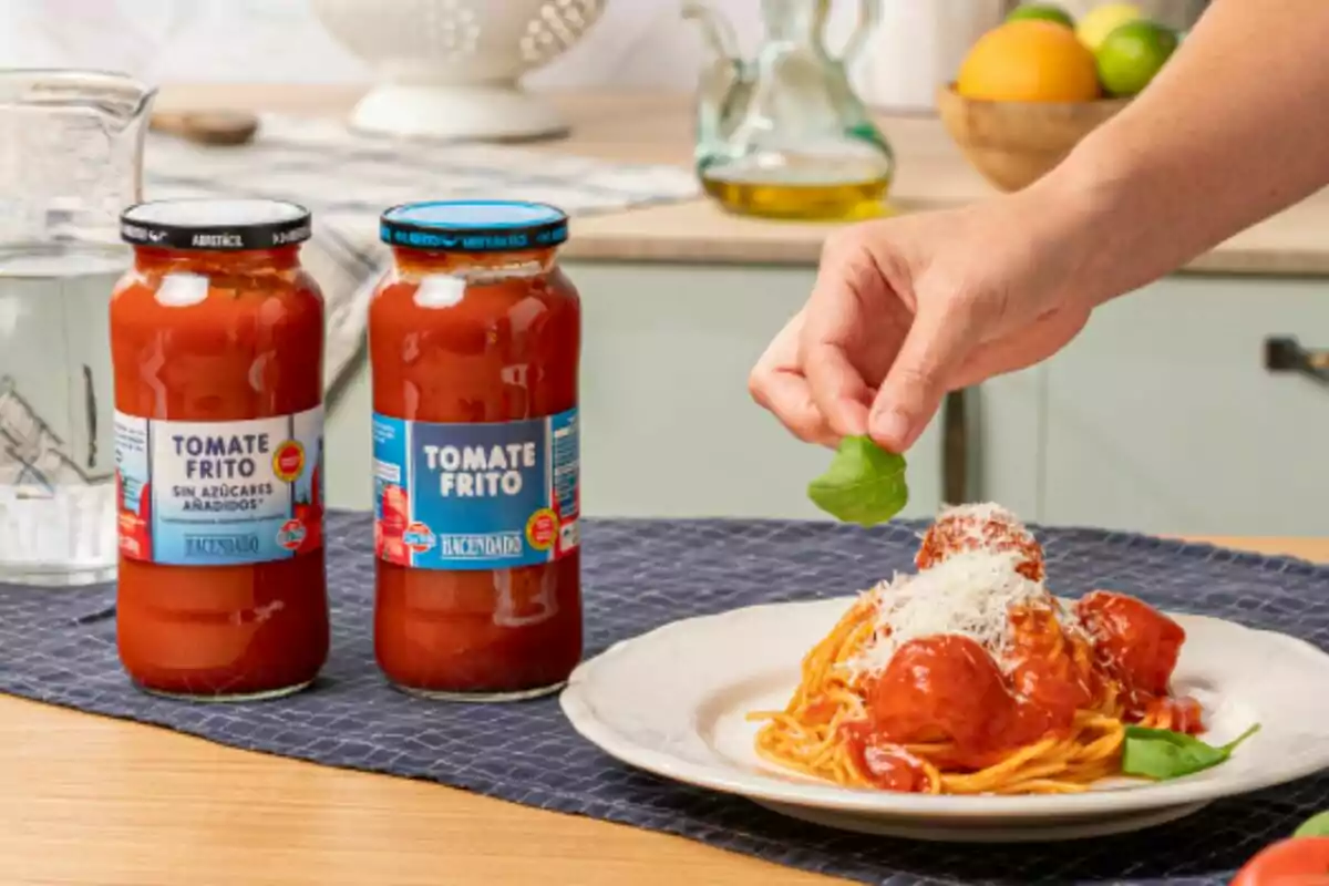 Dos frascos de tomate frito junto a un plato de espaguetis con albóndigas y una mano agregando una hoja de albahaca.
