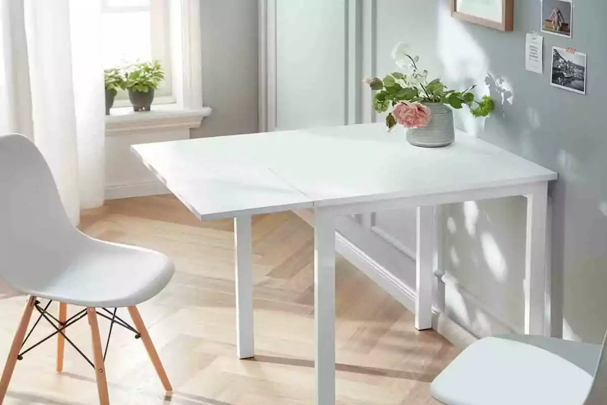 Una taula blanca amb una cadira blanca en una habitació lluminosa amb pis de fusta, una finestra amb cortines blanques i plantes a l'ampit, i una paret grisa amb decoracions i un gerro amb flors sobre la taula. Una taula blanca amb una cadira blanca en una habitació lluminosa amb pis de fusta, una finestra amb cortines blanques i plantes a l'ampit, i una paret grisa amb decoracions i un gerro amb flors sobre la taula.