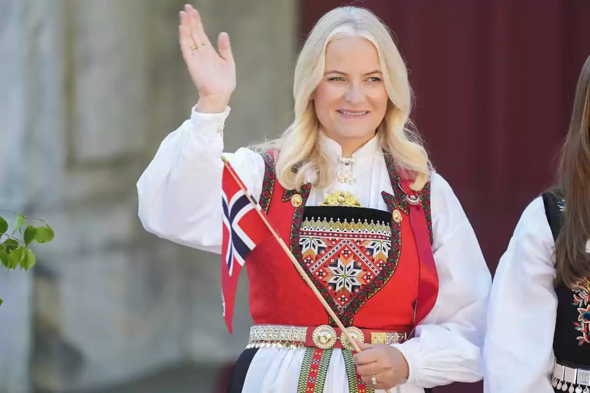 Mette-Marit amb un vestit tradicional noruec sosté una bandera mentre somriu i saluda. Mette-Marit amb un vestit tradicional noruec sosté una bandera mentre somriu i saluda.