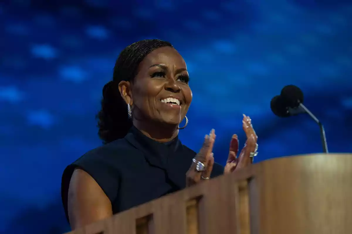 Michelle Obama smiling and clapping behind a podium with a blue background and a microphone to the side Michelle Obama smiling and clapping behind a podium with a blue background and a microphone to the side