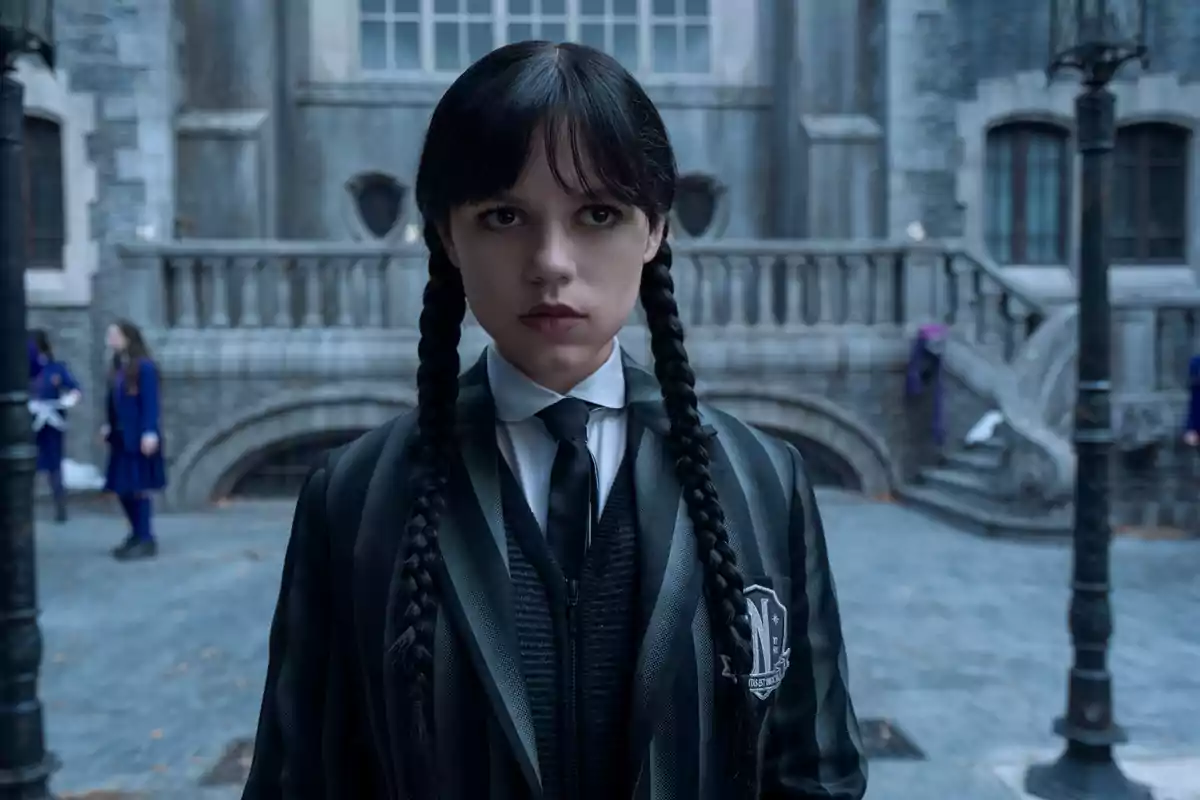 Jenna Ortega with braids and a serious expression stands in front of a Gothic-style building, wearing a dark uniform with a tie in Wednesday.