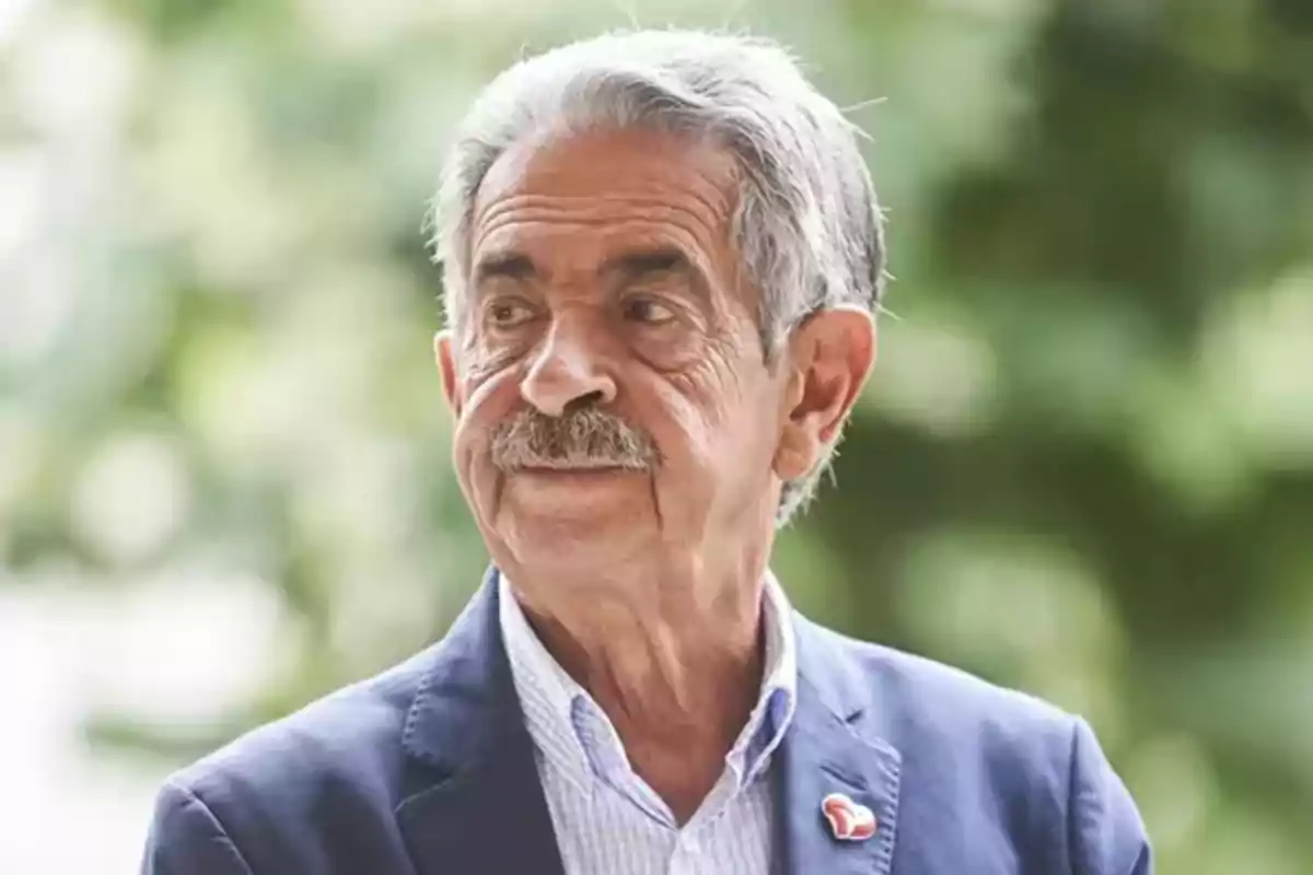 Miguel Ángel Revilla amb bigoti i cabell canós porta un vestit blau i una camisa de quadres mentre mira cap a un costat amb un fons desenfocat de vegetació.