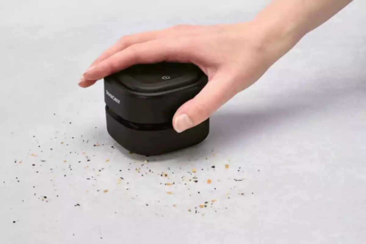 Una mà utilitzant un petit aspirador de taula negre per recollir molles sobre una superfície grisa. Una mà utilitzant un petit aspirador de taula negre per recollir molles sobre una superfície grisa.