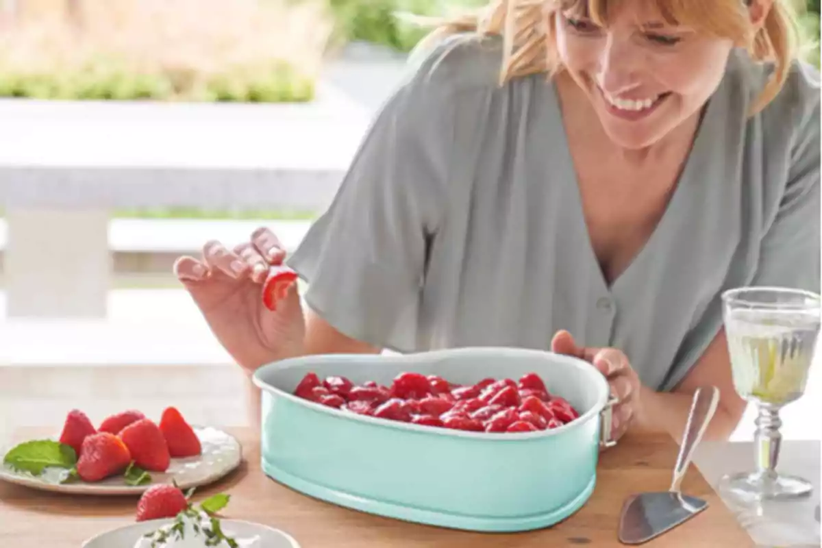 Una dona somrient prepara unes postres amb maduixes en un recipient blau mentre està asseguda en una taula a l'aire lliure.