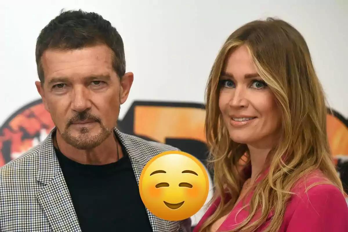Antonio Banderas and Nicole Kimpel pose together in front of a white wall with a smiling emoji superimposed in the center of the image.