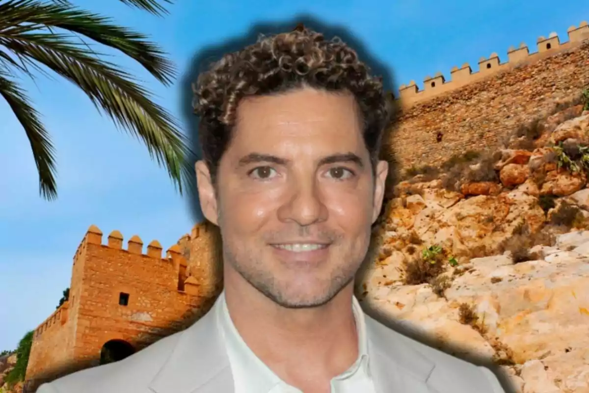 David Bisbal amb cabell arrissat i una jaqueta clara somriu davant d'un fons d'una fortalesa de pedra i una palmera sota un cel blau.