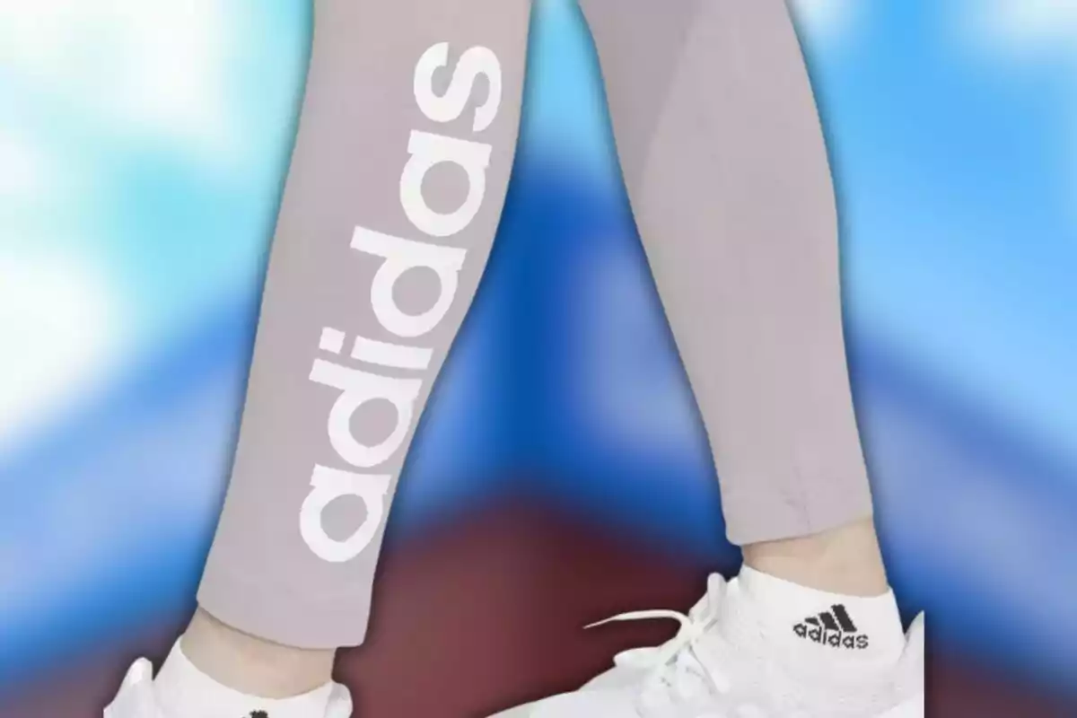 Persona vestint leggings grisos amb el logotip de Adidas i sabatilles blanques de la mateixa marca sobre un fons desenfocat blau i vermell.