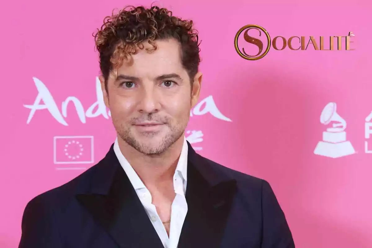 David Bisbal, vestit amb un vestit fosc i camisa blanca, posant davant d'un fons rosa amb logotips i text.