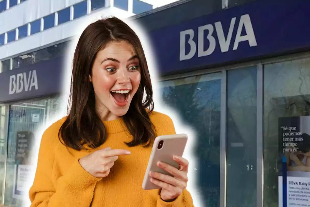 Una dona amb suèter groc mira emocionada el telèfon davant una sucursal de BBVA.