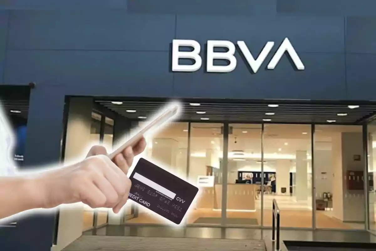 Una persona sosté un telèfon mòbil i una targeta de crèdit davant d'una sucursal del banc BBVA.