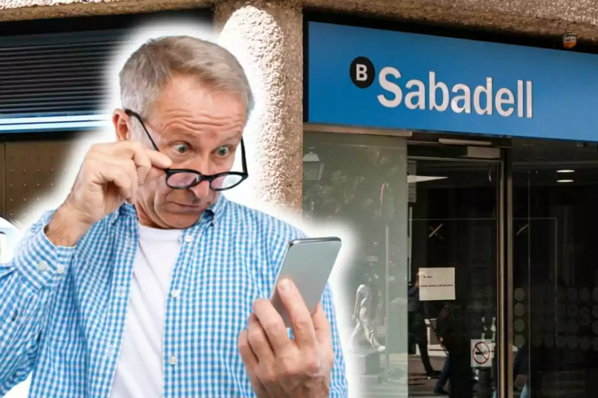 Un home gran amb una camisa a quadres blaus i ulleres mira sorprès el telèfon mòbil davant d'una sucursal del banc Sabadell.