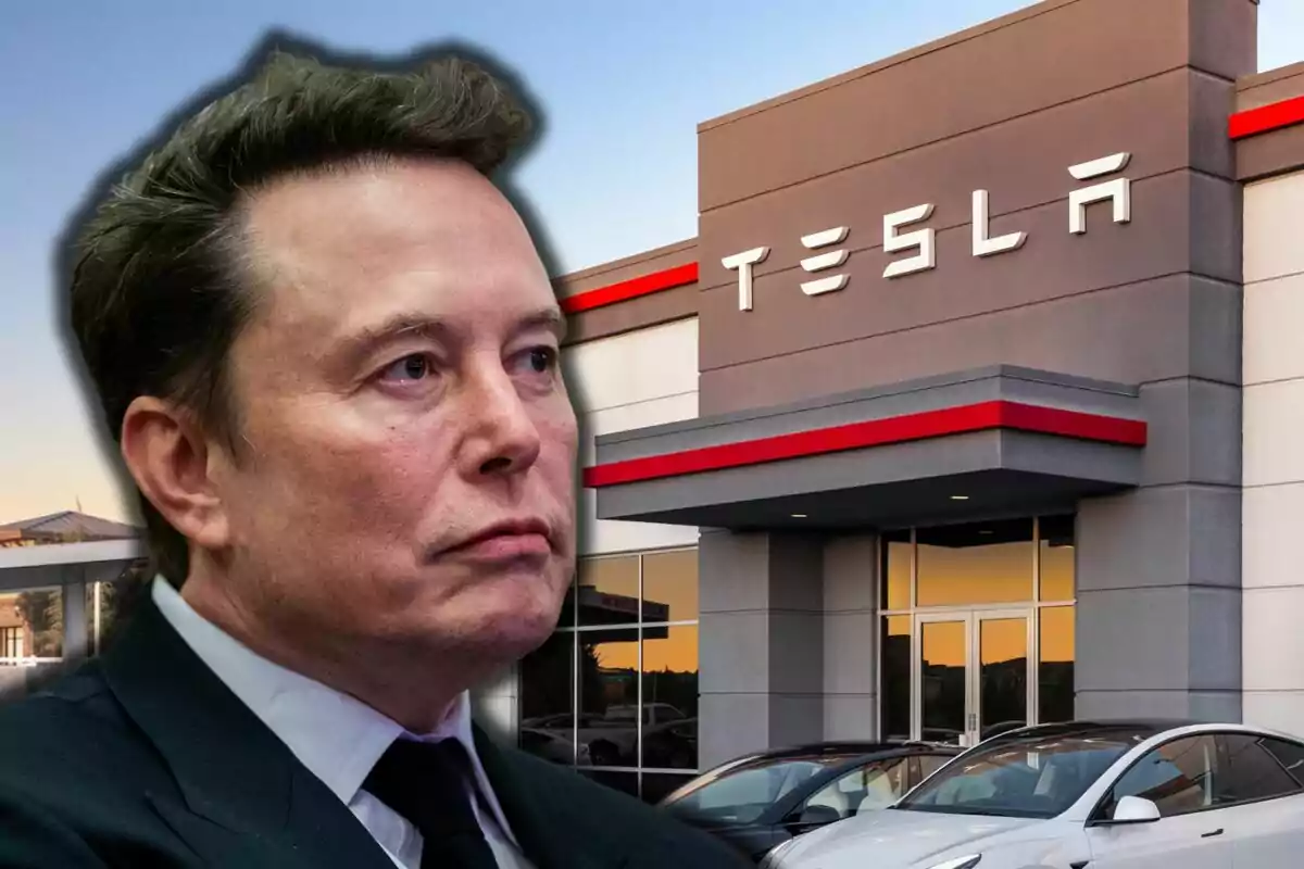 A man in a dark suit in front of a Tesla building with parked cars.