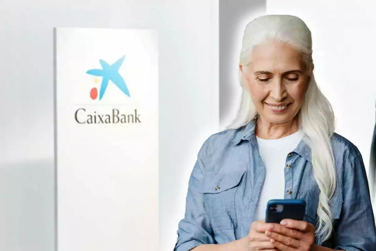 Una dona gran amb cabell blanc somriu mentre mira el telèfon mòbil, amb un cartell de CaixaBank al fons.