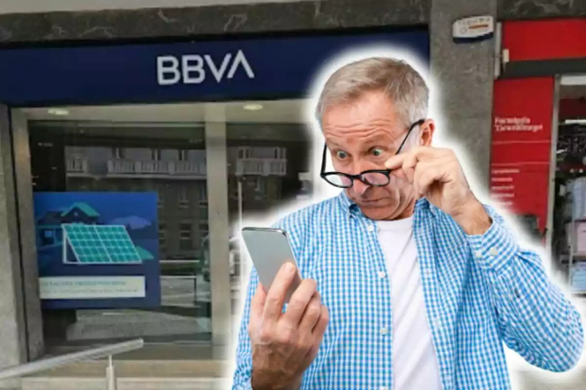 Un home gran amb camisa a quadres blaus i ulleres observa sorprès el telèfon mòbil davant d'una sucursal del banc BBVA.