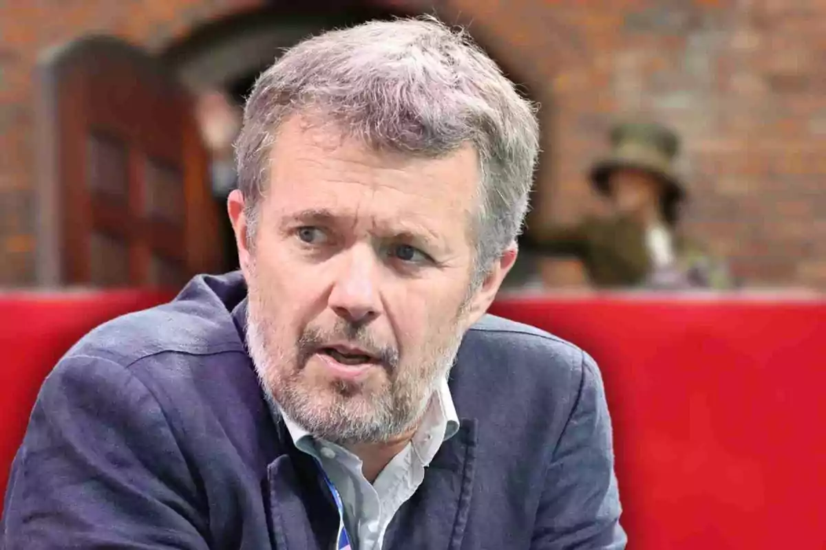 Frederic de Dinamarca amb barba curta i expressió seriosa assegut davant d’un fons desenfocat de maons i una persona al fons