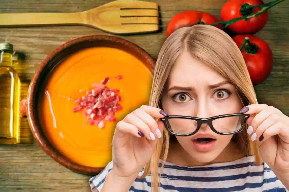 Una dona amb expressió de sorpresa sosté les seves ulleres davant d'un plat de sopa de tomàquet amb trossos de pernil, acompanyat d'una ampolla d'oli, una cullera de fusta i tomàquets frescos sobre una taula de fusta.