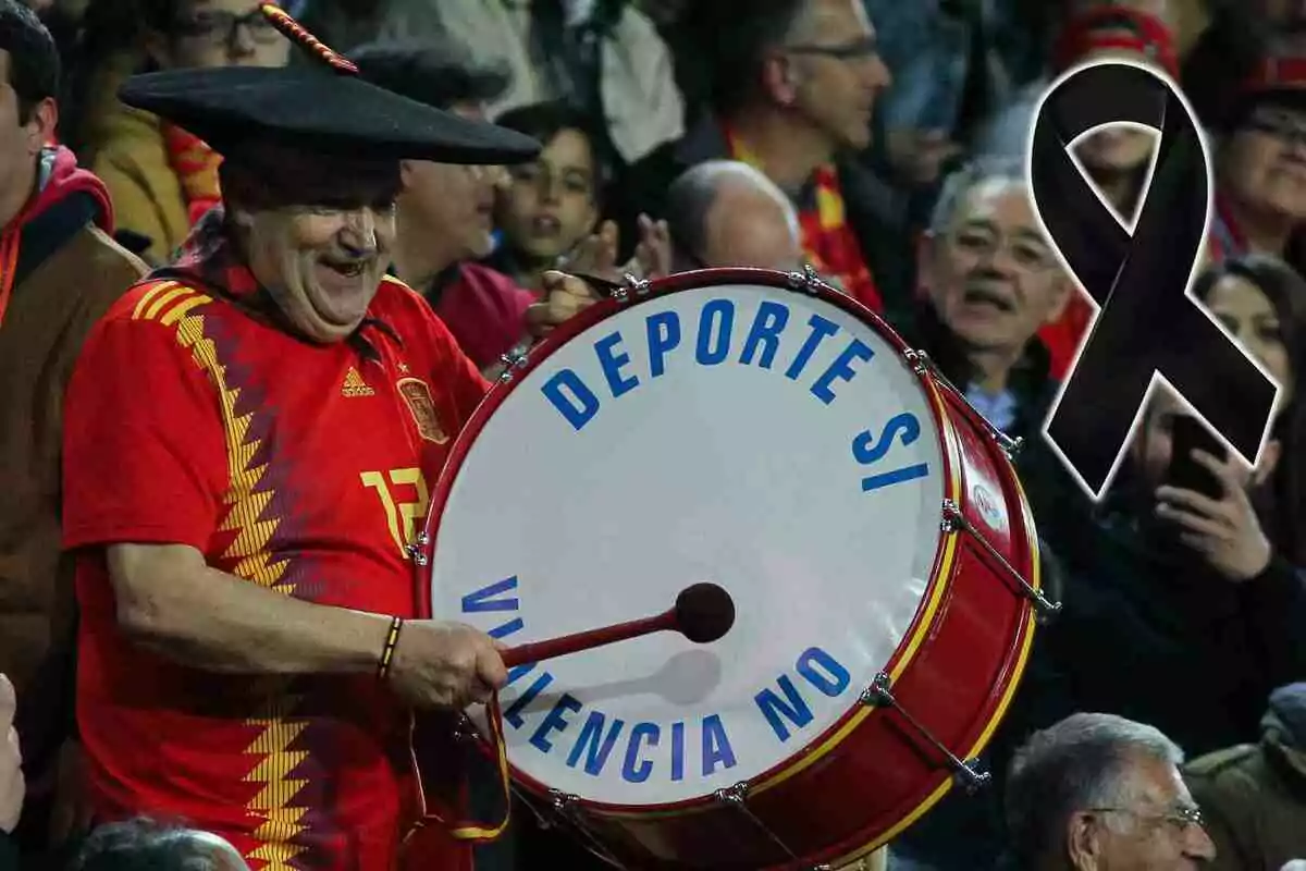 Manolo El del Bombo amb samarreta vermella i barret negre toca un tambor que diu "Esport sí, València no" mentre un llaç negre apareix al costat.