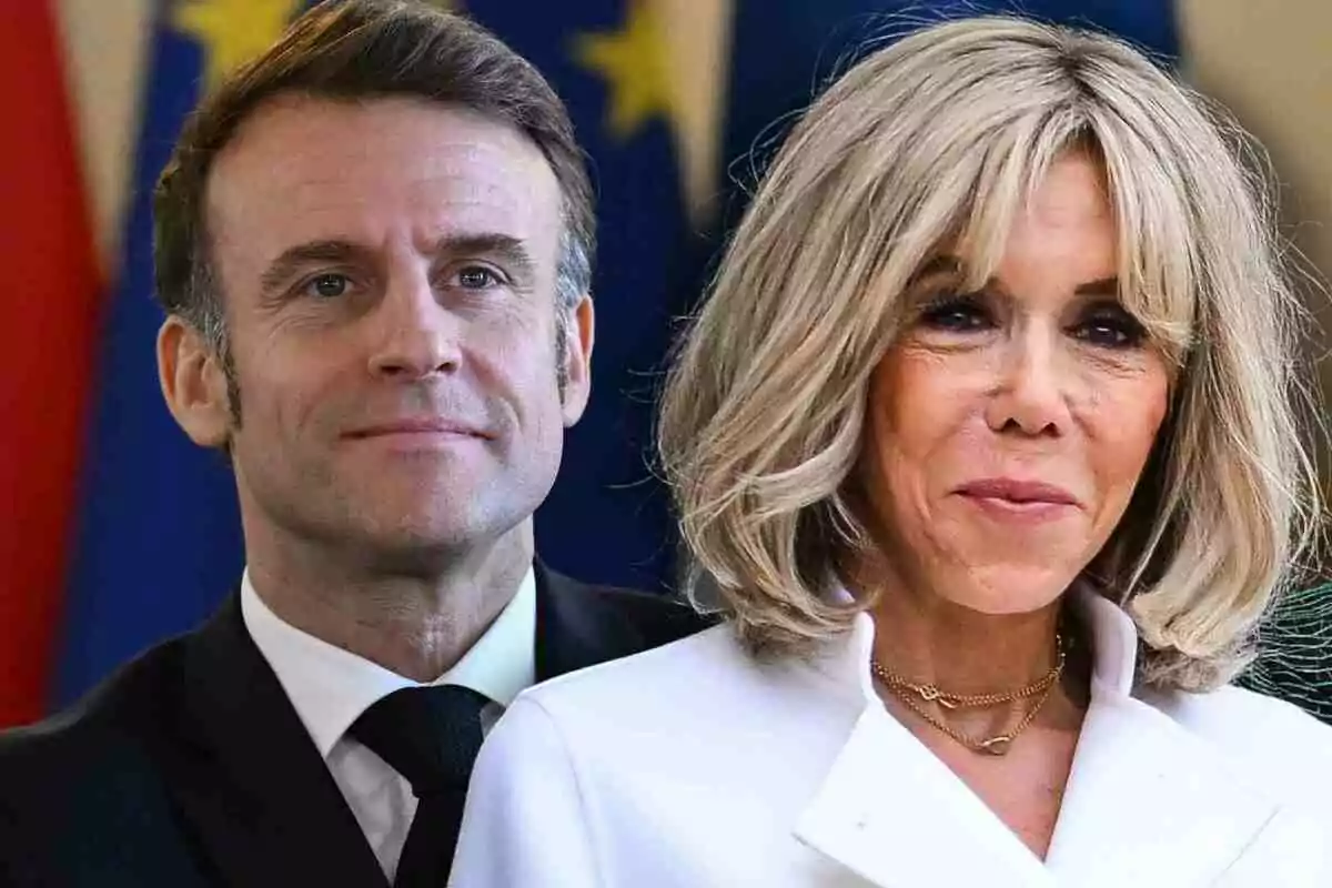 Emmanuel Macron in a dark suit and Brigitte Macron with blonde hair and a white coat pose together in front of a background with flags.