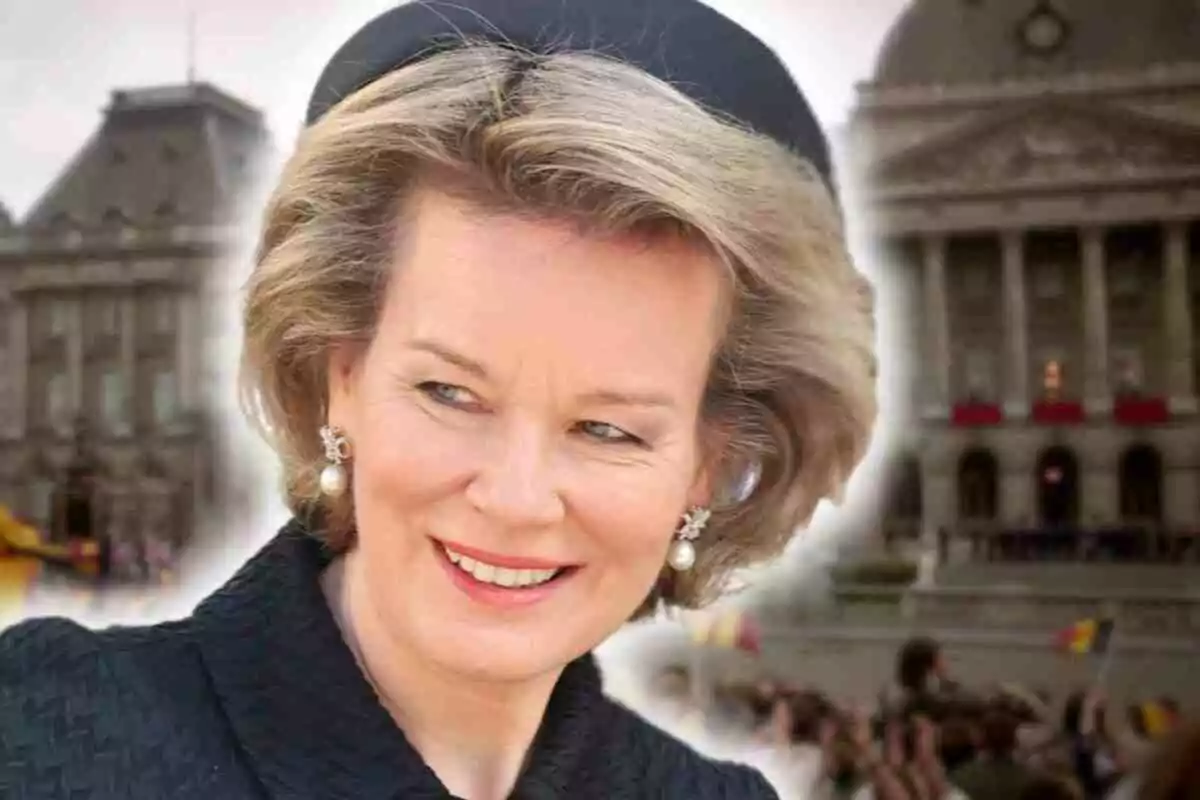 Mathilde of Belgium with pearl earrings and a black dress smiles in front of a historic building with people and flags in the background