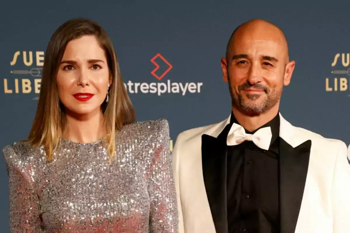 Natalia Sánchez amb vestit platejat brillant i Alain Hernández amb esmòquing blanc i negre posen junts en un esdeveniment amb fons blau i logotips.