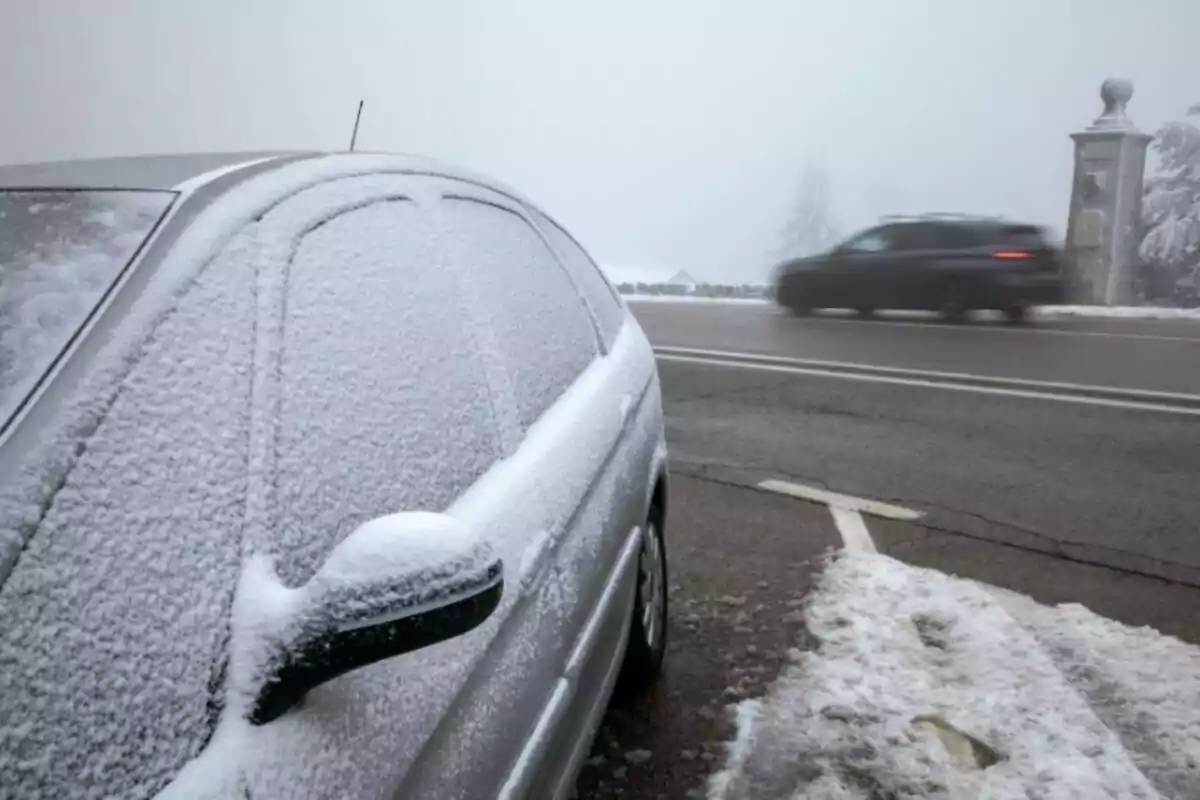 Un cotxe cobert de neu estacionat al costat d?una carretera mentre un altre vehicle passa ràpidament en un dia ennuvolat.