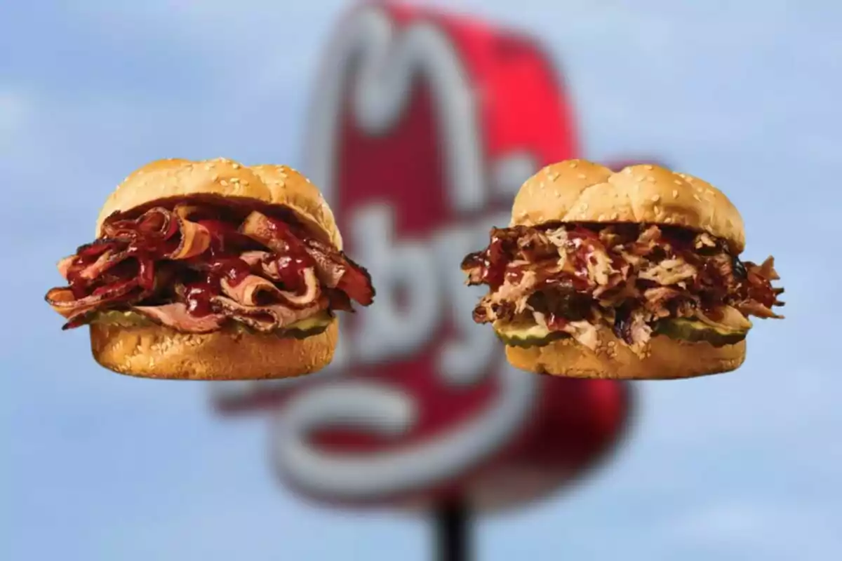 Dos sándwiches de carne con salsa barbacoa y pepinillos frente a un cartel borroso de un restaurante de comida rápida. Dos sándwiches de carne con salsa barbacoa y pepinillos frente a un cartel borroso de un restaurante de comida rápida.