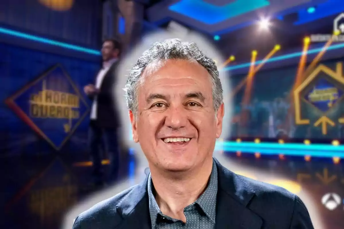 Roberto Brasero somrient amb cabell canós i arrissat, vestit amb una jaqueta fosca i una camisa blava, apareix en primer pla amb un fons borrós del set d''El Hormiguero' il·luminat amb llums blaves i grogues.