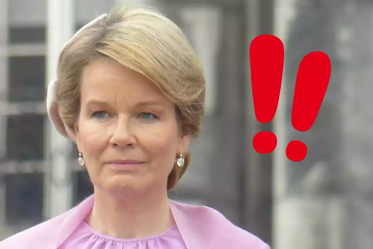 Mathilde of Belgium with a serious expression, dressed in pink, with red exclamation marks beside her.