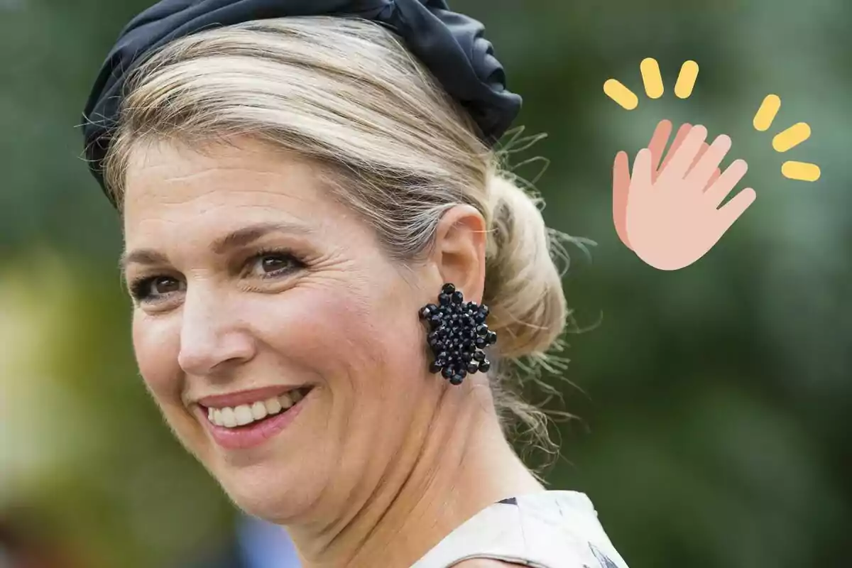 La reina Màxima d'Holanda somrient amb un pentinat recollit i un accessori negre al cabell porta una arracada gran i fosca mentre un emoji d'aplaudiment apareix al costat.