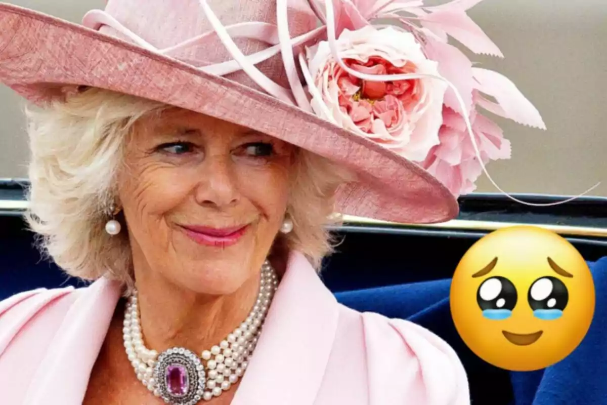 Queen Camila, wearing an elegant pink hat adorned with flowers and a pearl necklace, smiles while sitting in a vehicle, accompanied by an emoji with bright eyes and tears of emotion.