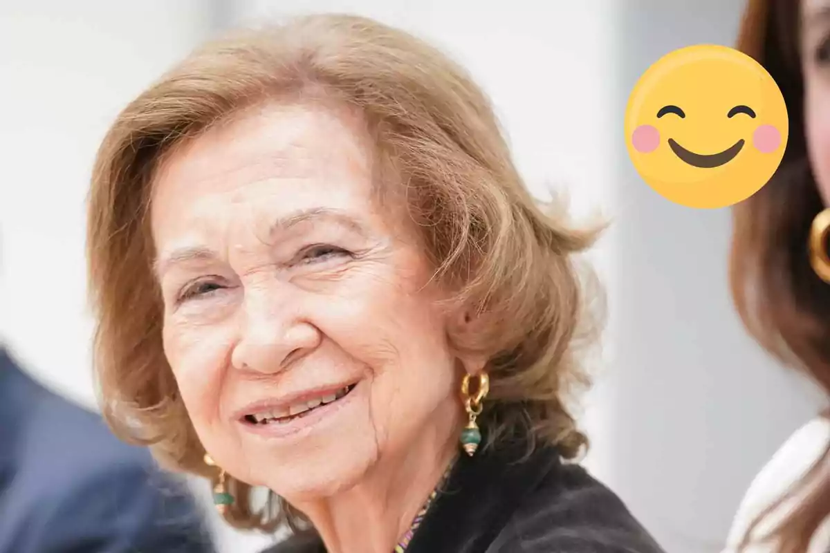 La reina Sofia somrient amb cabell castany clar i pendents daurats al costat d'un emoji somrient.