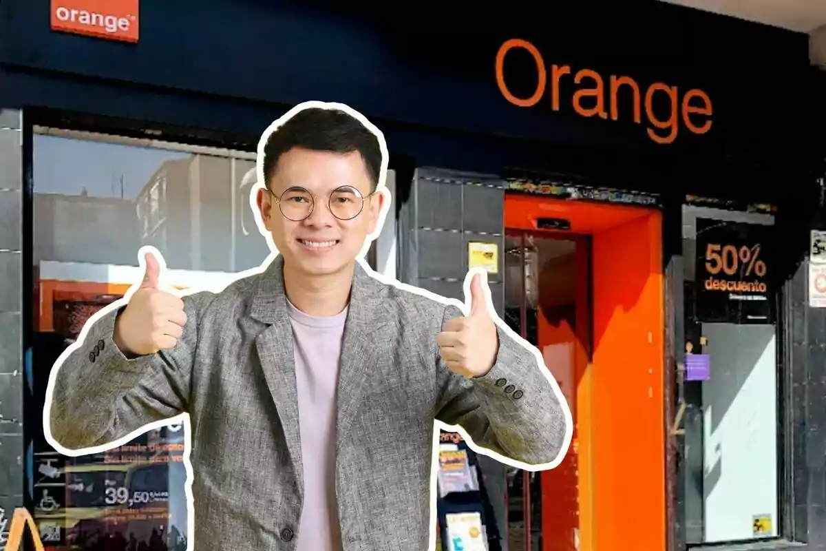 Hombre joven con gafas y saco gris sonriendo y levantando ambos pulgares frente a una tienda de Orange