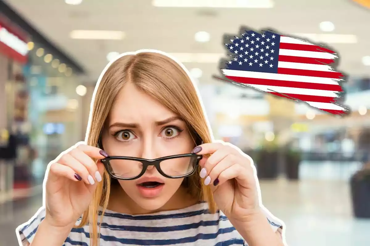 Una mujer con expresión de sorpresa sostiene sus gafas en un entorno interior con una bandera de Estados Unidos en el fondo.