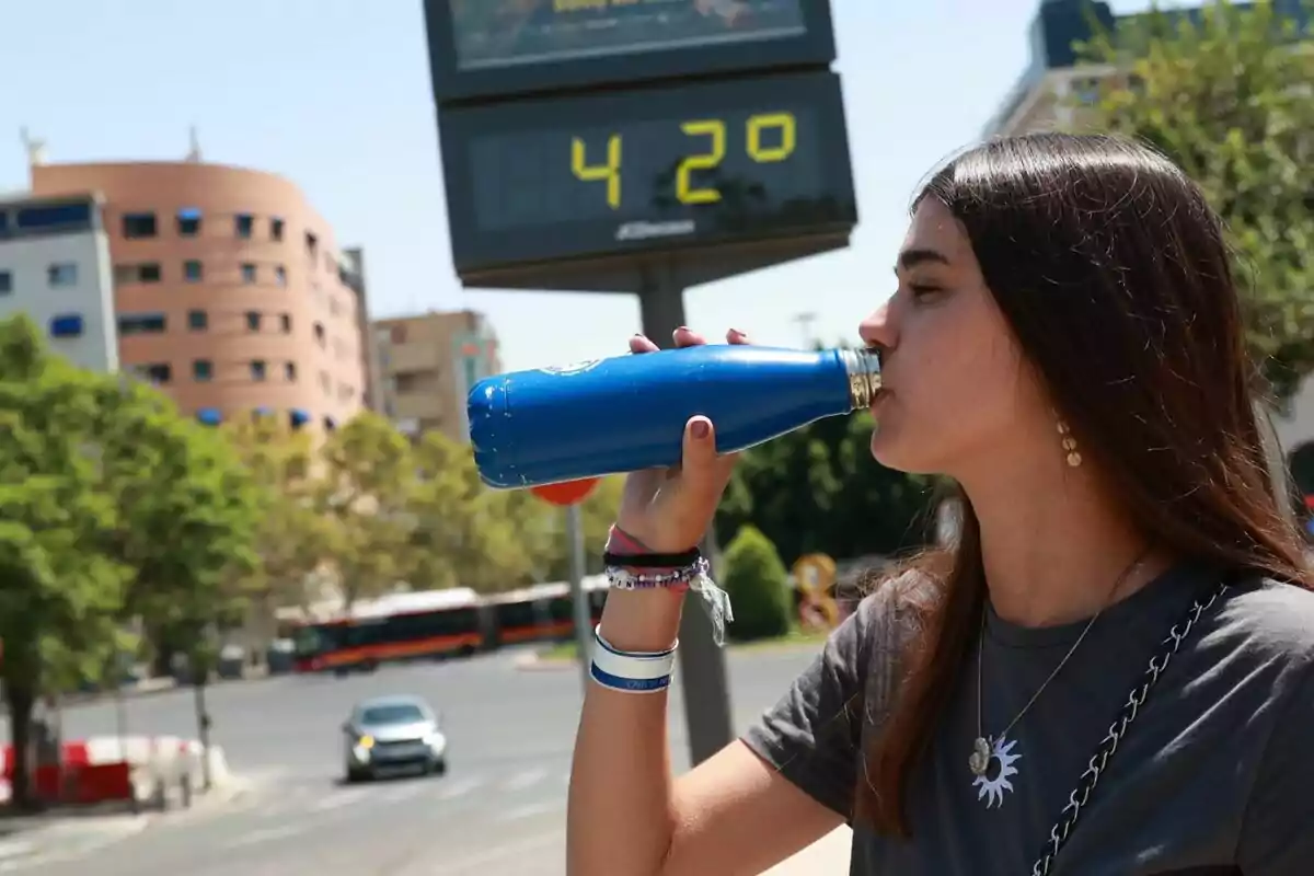 Jove bevent aigua d’una ampolla blava al carrer durant un dia calorós amb un termòmetre que marca 42 graus al fons.