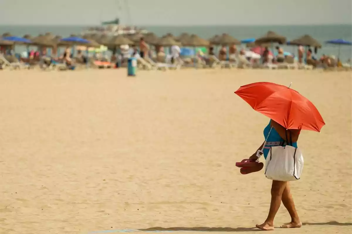 Dona caminant descalça per la platja amb les xancletes a la mà i un para-sol taronja