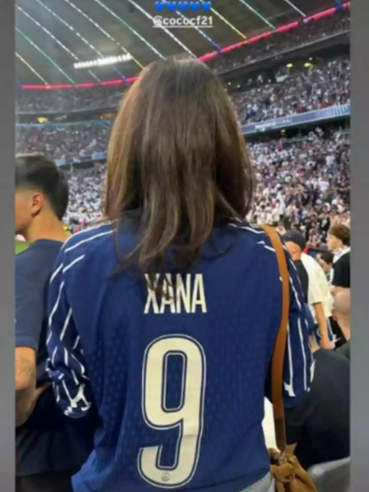 Dona d’esquena amb samarreta blava de futbol que porta el nom Xana i el número 9 en un estadi ple d’espectadors Dona d’esquena amb samarreta blava de futbol que porta el nom Xana i el número 9 en un estadi ple d’espectadors