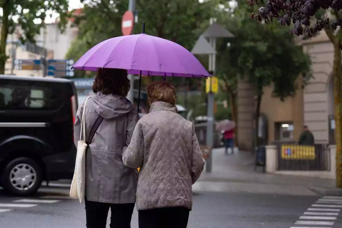 Dues dones d'esquena caminant pel carrer amb jaqueta i un paraigua lila