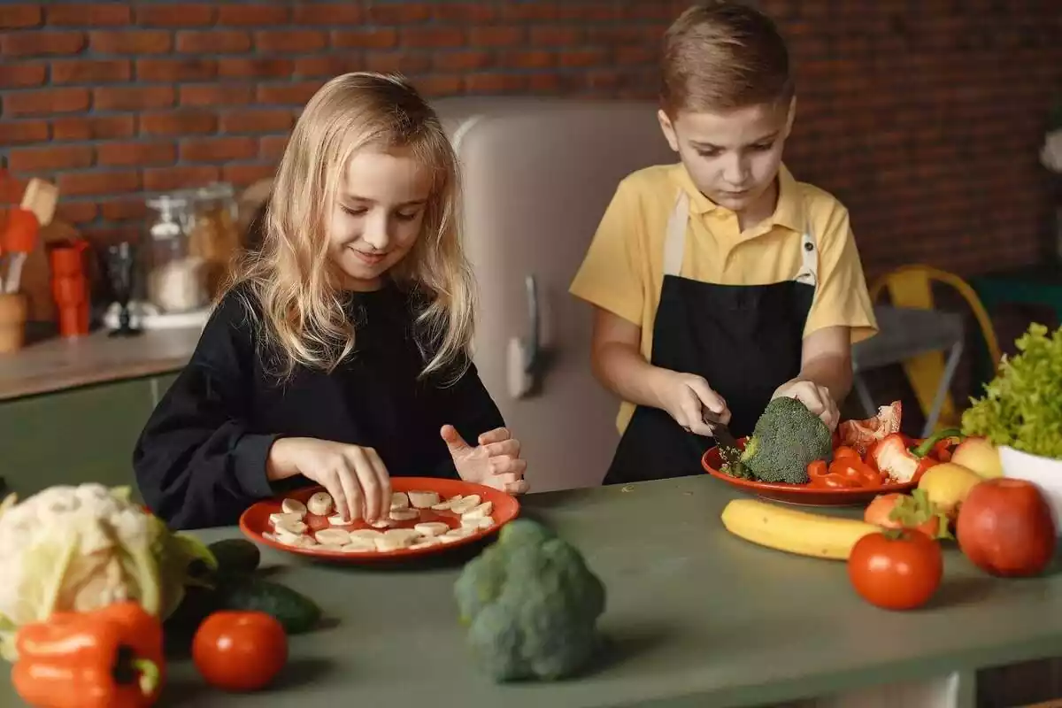 Dos nens cuinant amb diverses fruites i verdures com plàtan i bròquil a la cuina Dos nens cuinant amb diverses fruites i verdures com plàtan i bròquil a la cuina