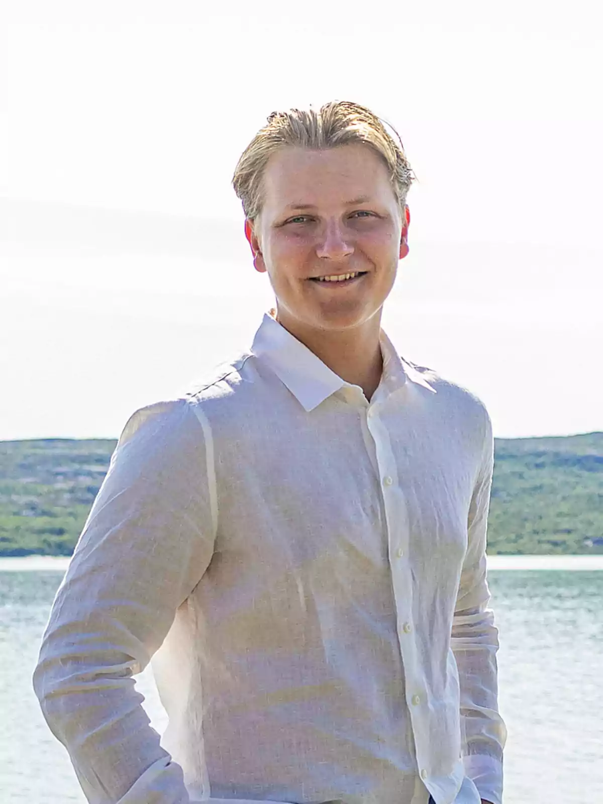 Sverre de Noruega amb camisa blanca somriu davant d'un paisatge d'aigua i turons. Sverre de Noruega amb camisa blanca somriu davant d'un paisatge d'aigua i turons.