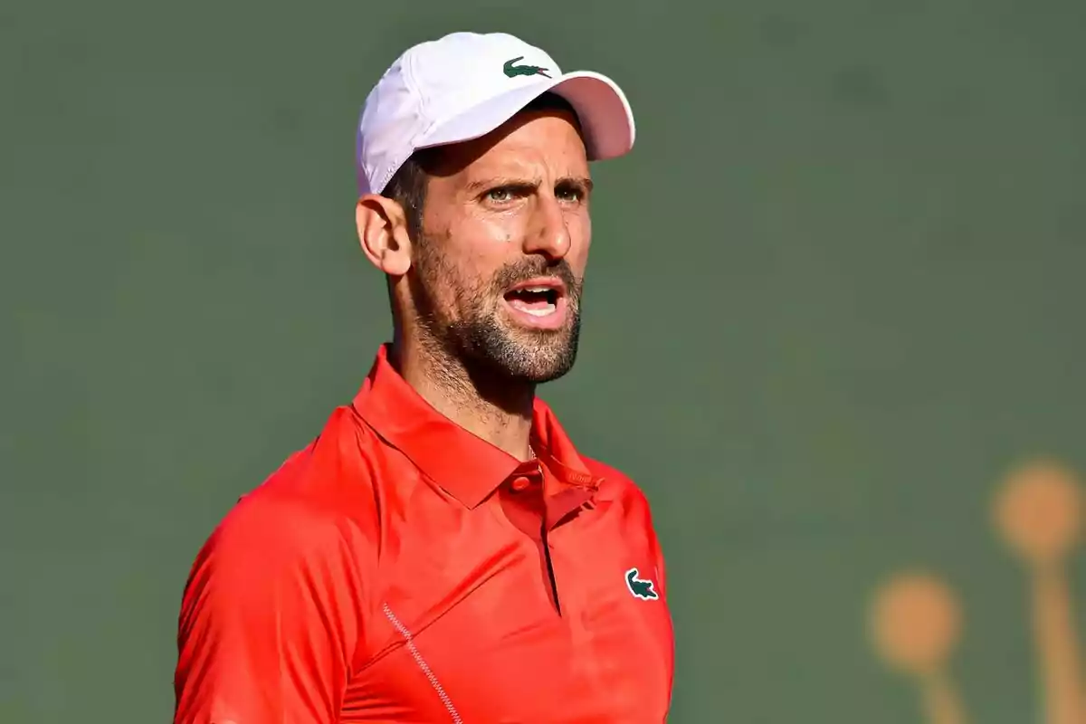 Novak Djokovic con camiseta roja y gorra blanca en una cancha de tenis. Novak Djokovic con camiseta roja y gorra blanca en una cancha de tenis.