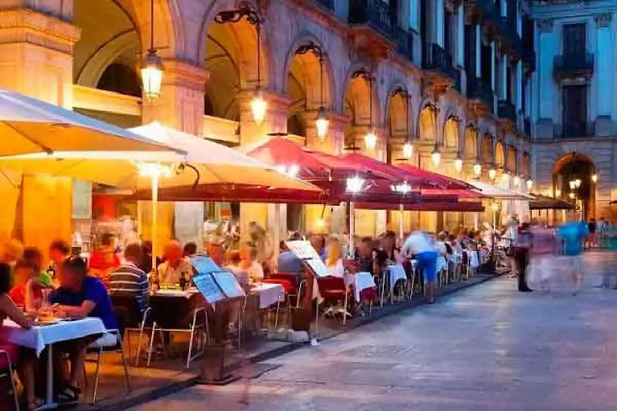 Persones sopant a l'aire lliure en una terrassa il·luminada a la nit en una plaça amb arcs i fanals.