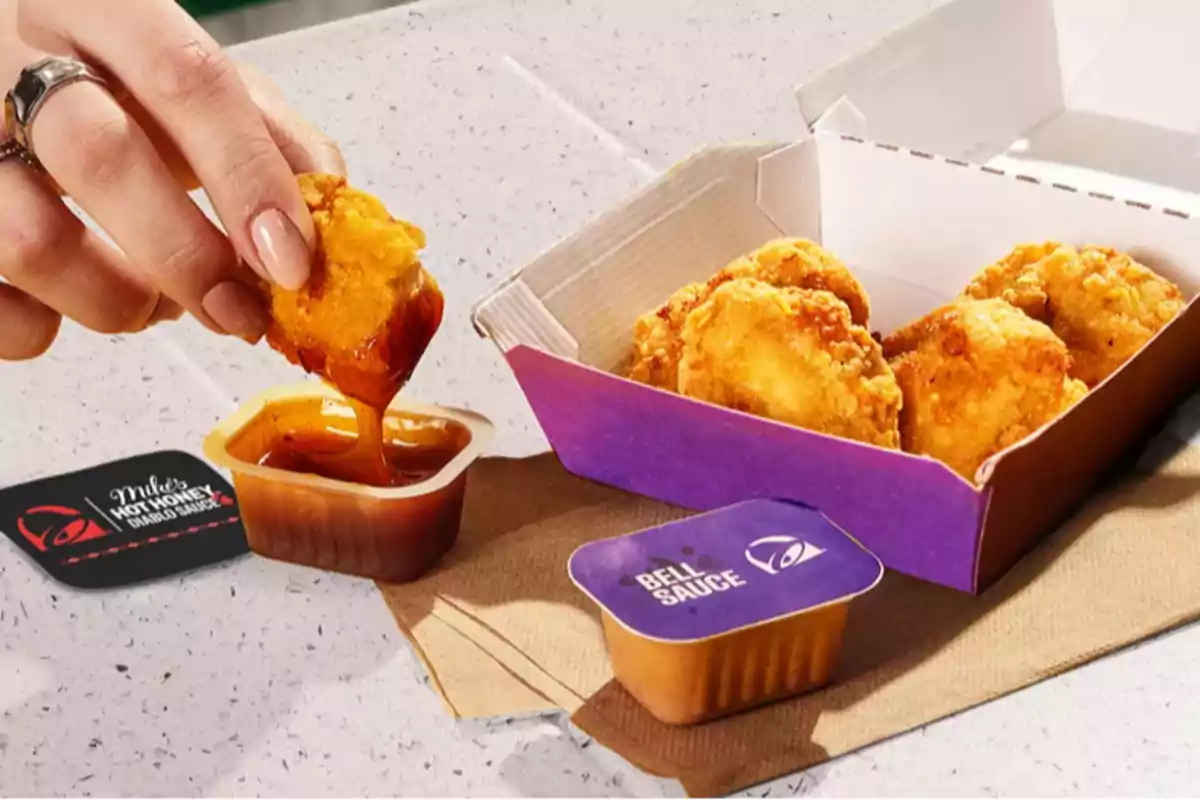 Una mà submergint un tros de pollastre arrebossat en una salsa, amb una caixa de nuggets i dos recipients de salsa sobre una taula. Una mà submergint un tros de pollastre arrebossat en una salsa, amb una caixa de nuggets i dos recipients de salsa sobre una taula.
