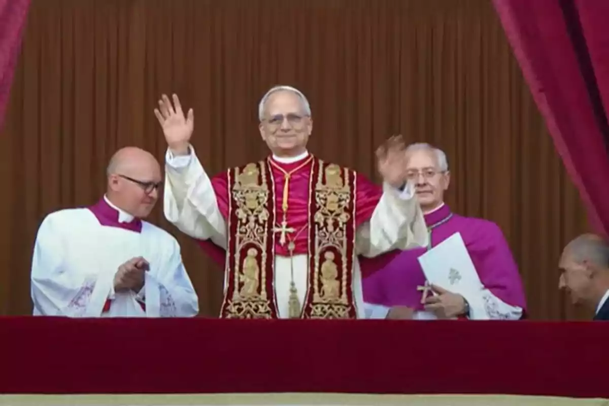 Un home vestit amb indumentària religiosa aixeca les mans mentre està dret en un balcó, acompanyat per altres dos homes amb vestimenta clerical.