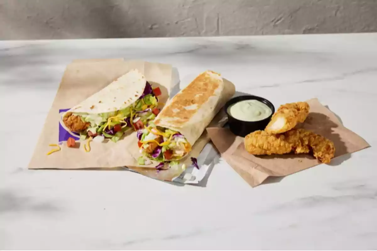 Tres piezas de pollo empanizado acompañadas de una salsa cremosa, un taco y un burrito rellenos de pollo, lechuga, jitomate y queso sobre una mesa blanca Tres piezas de pollo empanizado acompañadas de una salsa cremosa, un taco y un burrito rellenos de pollo, lechuga, jitomate y queso sobre una mesa blanca