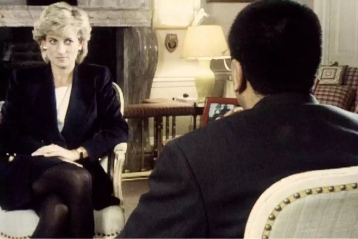 Lady Di asseguda en una cadira, vestida amb un vestit fosc, sembla estar en una entrevista amb un home d'esquena, en una habitació amb una làmpada i una xemeneia al fons. Lady Di asseguda en una cadira, vestida amb un vestit fosc, sembla estar en una entrevista amb un home d'esquena, en una habitació amb una làmpada i una xemeneia al fons.