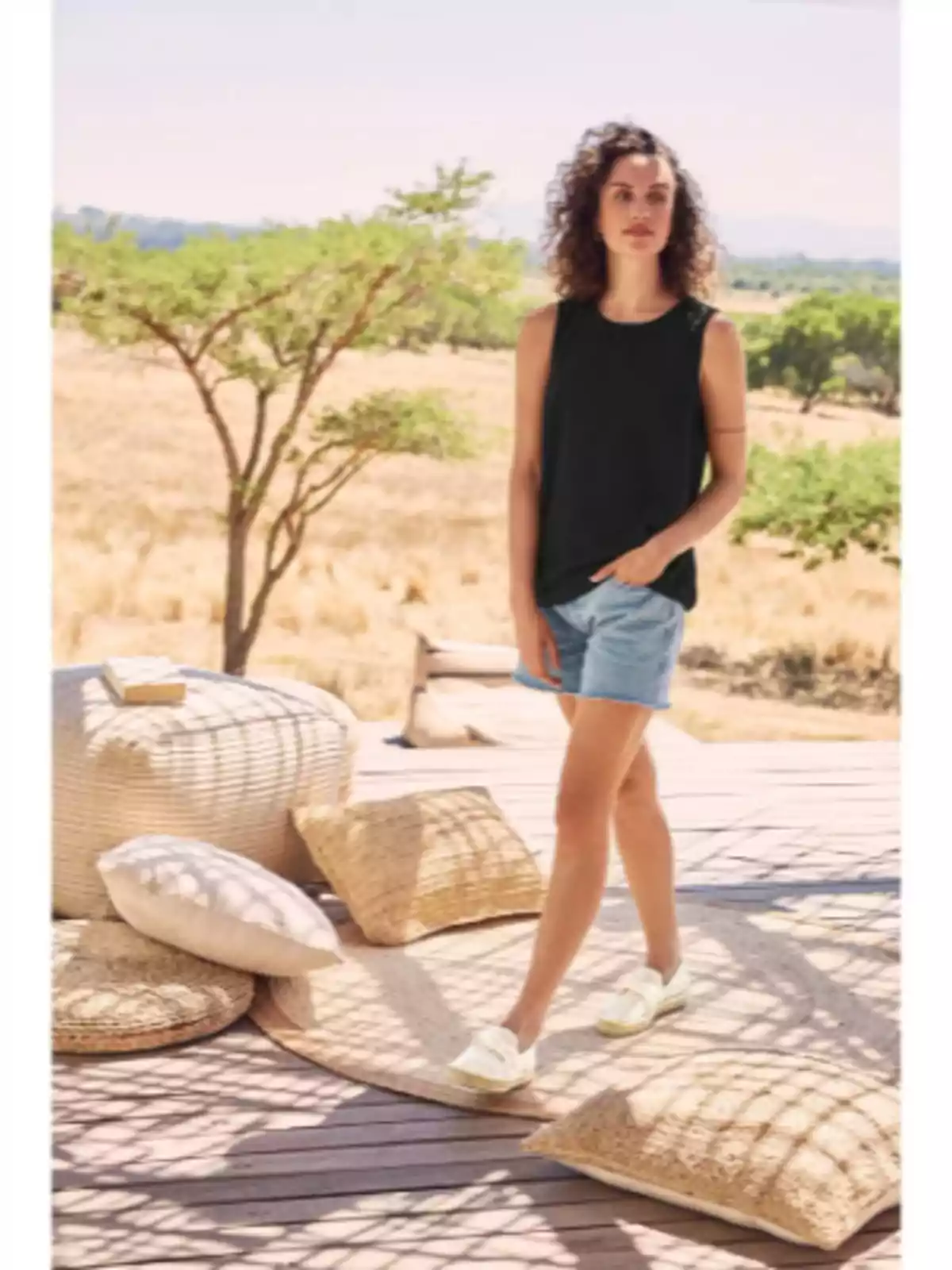 Dona de cabells arrissats amb samarreta negra i pantalons curts de mezclilla caminant per una terrassa de fusta decorada amb coixins i pufs en un entorn natural i assolellat. Dona de cabells arrissats amb samarreta negra i pantalons curts de mezclilla caminant per una terrassa de fusta decorada amb coixins i pufs en un entorn natural i assolellat.
