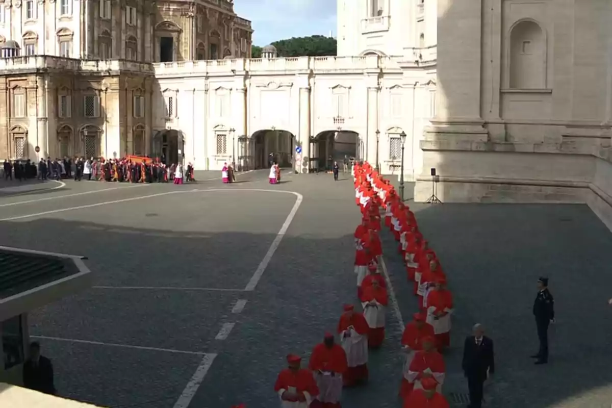 Un grup de cardenals vestits de vermell camina en fila per una plaça davant d'un edifici històric. Un grup de cardenals vestits de vermell camina en fila per una plaça davant d'un edifici històric.