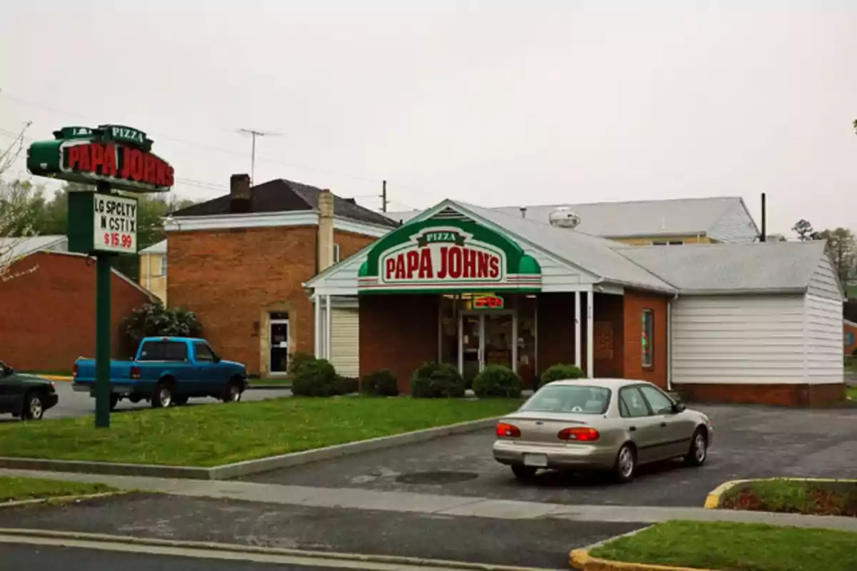 Facade of a Papa John’s pizzeria with a large sign and cars parked in front on a cloudy day Facade of a Papa John’s pizzeria with a large sign and cars parked in front on a cloudy day
